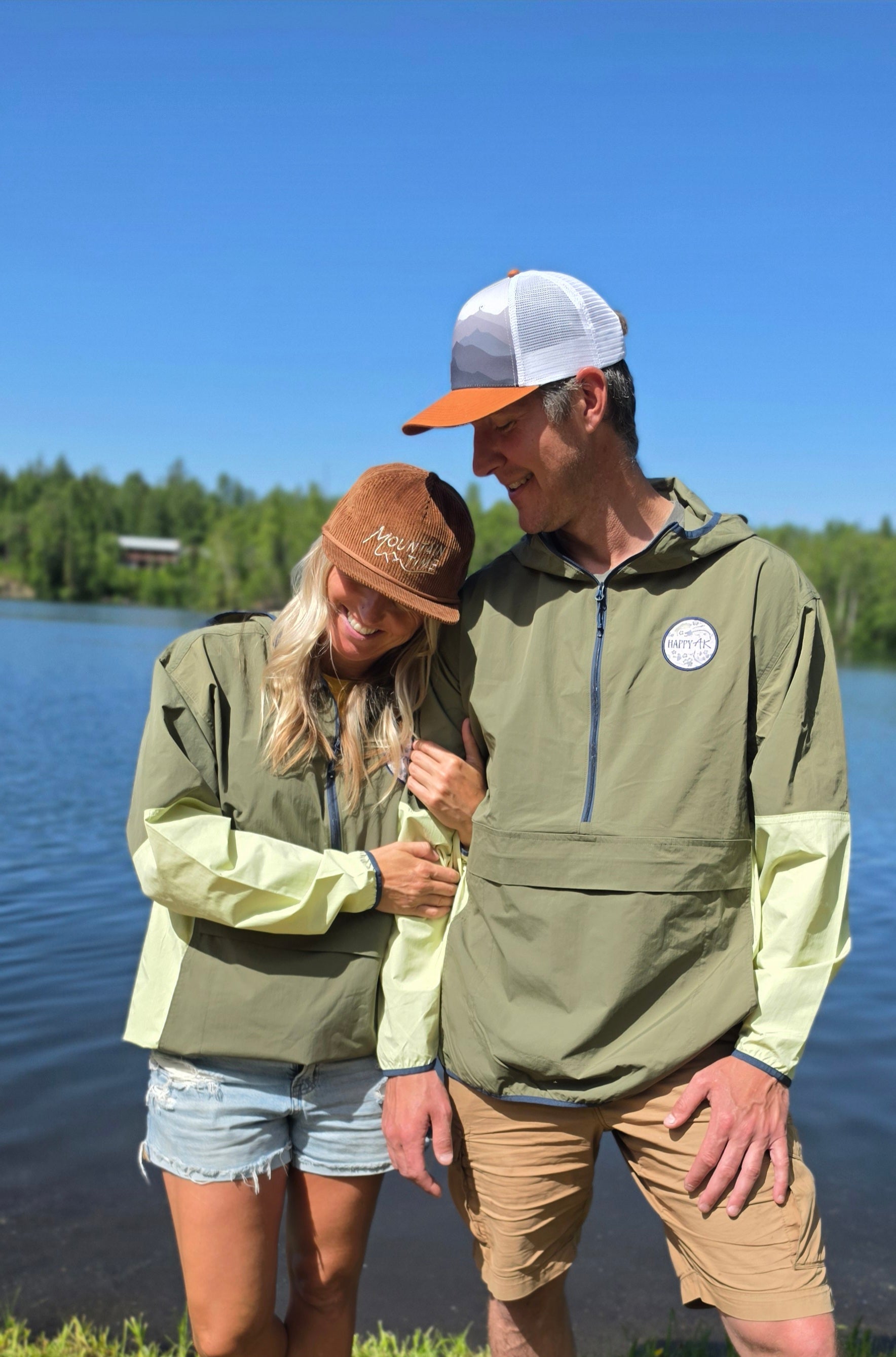 Two people wearing green jackets standing by a lake with trees in the background